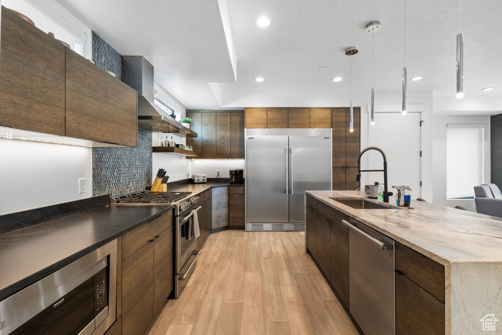 Kitchen featuring built in appliances, light wood finished floors, open shelves, decorative backsplash, and a large island with sink