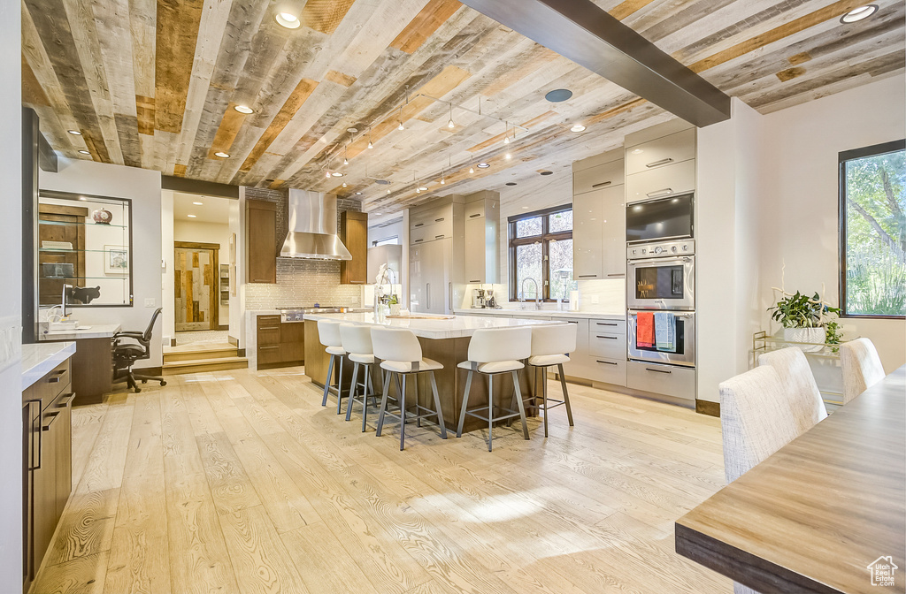 Kitchen featuring wall chimney exhaust hood, appliances with stainless steel finishes, a large island with sink, a breakfast bar, and light countertops