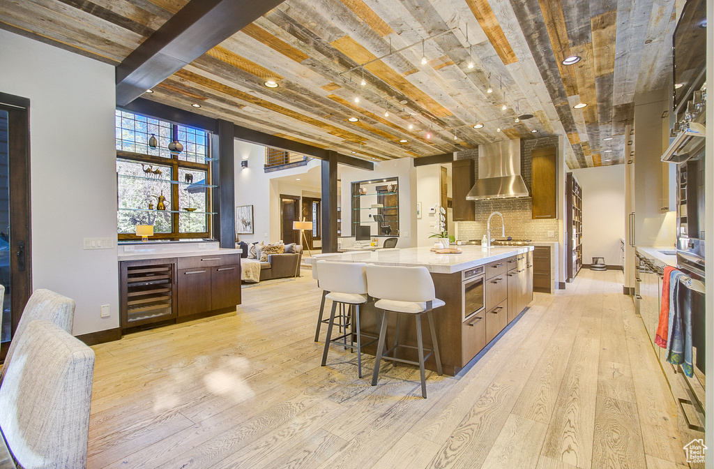 Kitchen featuring wall chimney exhaust hood, wine cooler, light wood finished floors, light countertops, and a kitchen bar