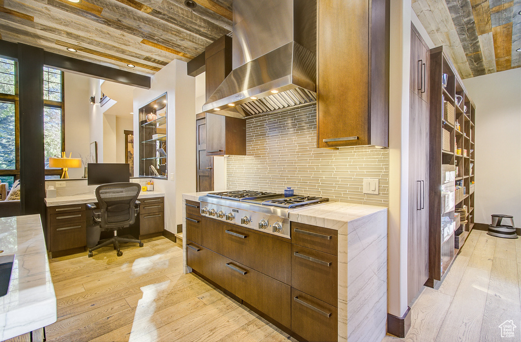 Kitchen with wall chimney range hood, stainless steel gas cooktop, light wood-style flooring, backsplash, and built in study area