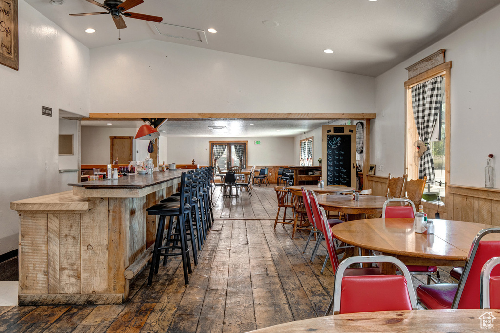 Dining space featuring wood-type flooring, a ceiling fan, high vaulted ceiling, a wainscoted wall, and a bar