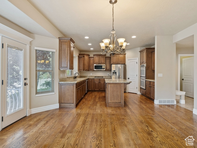 Kitchen with stainless steel appliances, a chandelier, a kitchen island, light stone countertops, and brown cabinets