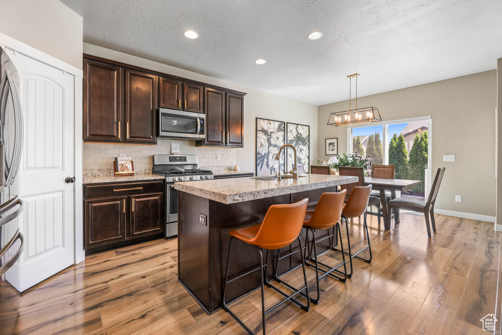 Kitchen featuring appliances with stainless steel finishes, light wood finished floors, a textured ceiling, decorative backsplash, and a center island with sink