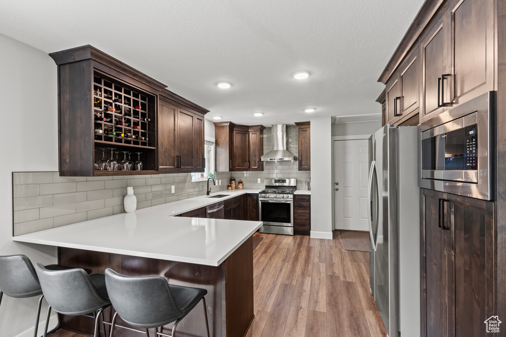Kitchen featuring stainless steel appliances, wall chimney exhaust hood, light wood-style flooring, a peninsula, and dark brown cabinets