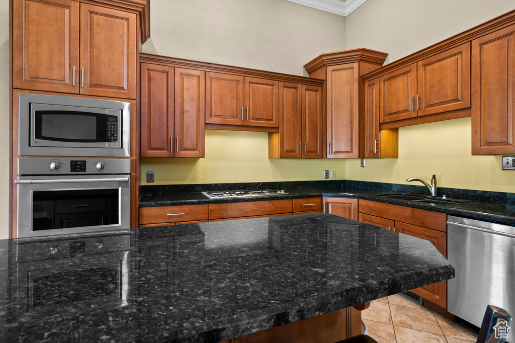 Kitchen with appliances with stainless steel finishes, ornamental molding, dark stone countertops, light tile patterned floors, and brown cabinetry