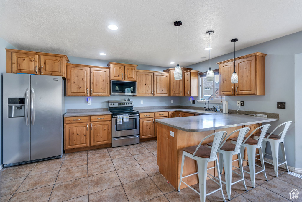 Kitchen featuring appliances with stainless steel finishes, a peninsula, pendant lighting, a kitchen bar, and brown cabinets