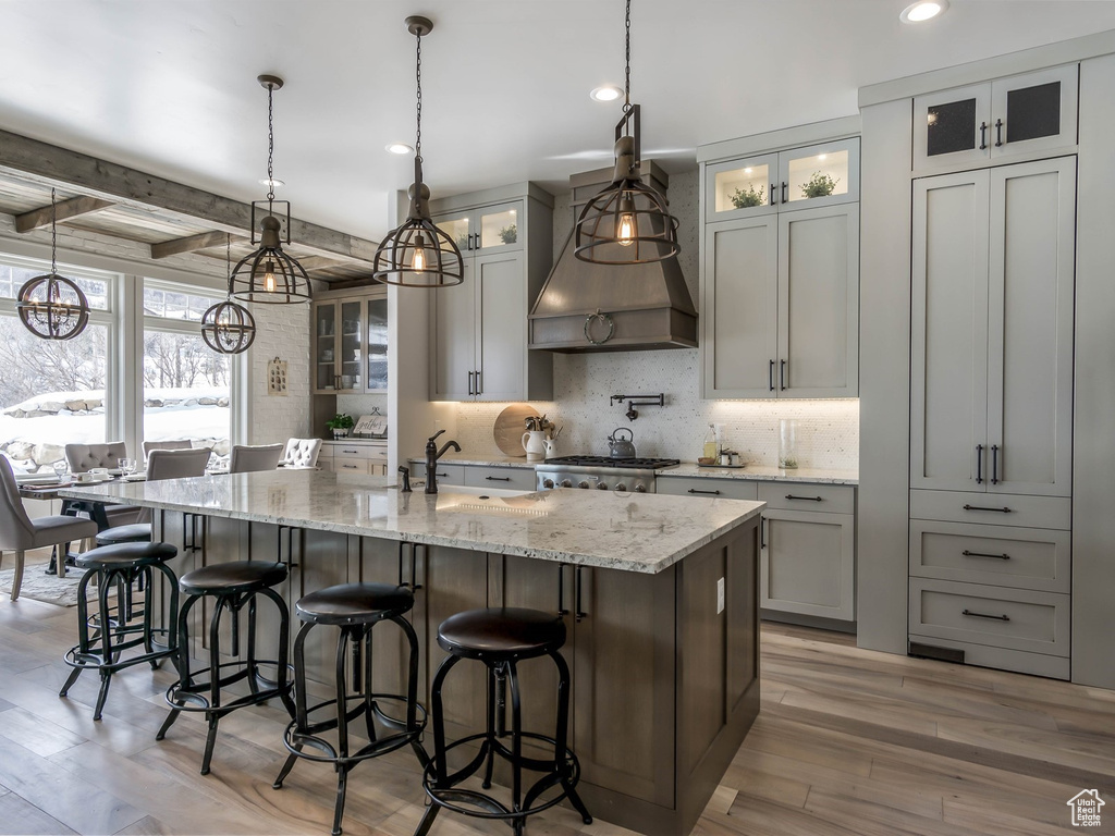 Kitchen featuring an island with sink, light wood finished floors, backsplash, light stone countertops, and decorative light fixtures