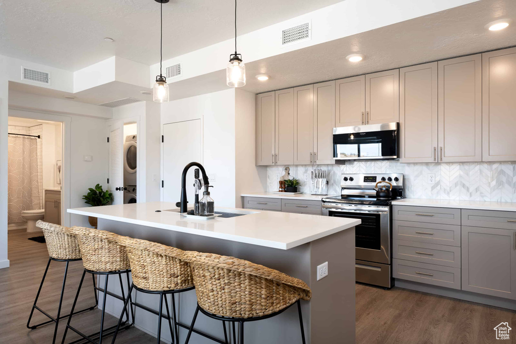 Kitchen featuring appliances with stainless steel finishes, stacked washing machine and dryer, gray cabinetry, light countertops, and an island with sink