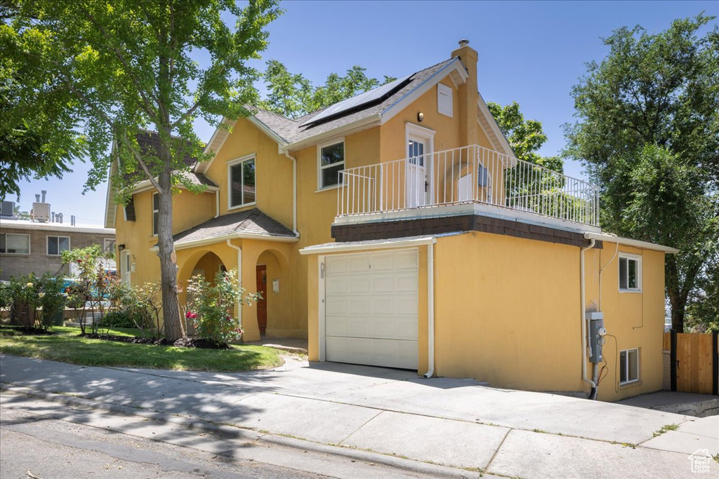 View of front facade featuring a balcony, an attached garage, roof mounted solar panels, stucco siding, and a chimney