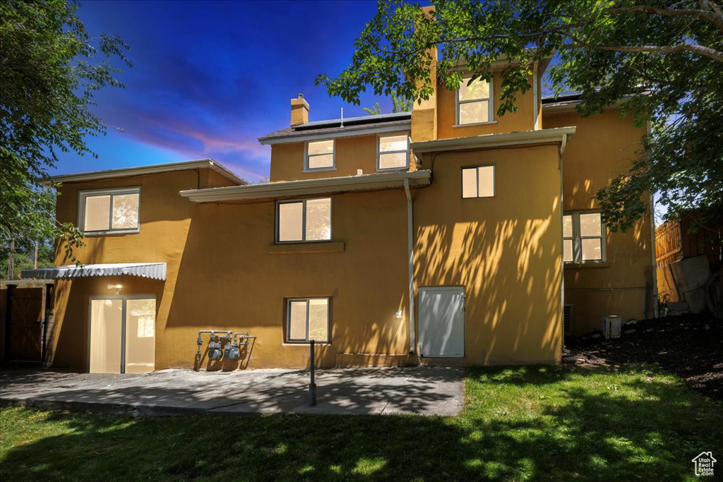 Back of house at dusk featuring stucco siding, a patio area, a chimney, roof mounted solar panels, and a lawn