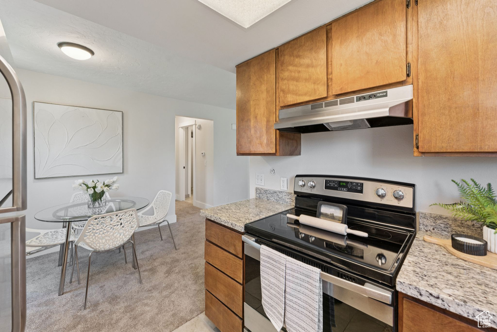 Kitchen with stainless steel electric range, under cabinet range hood, light countertops, brown cabinetry, and light carpet
