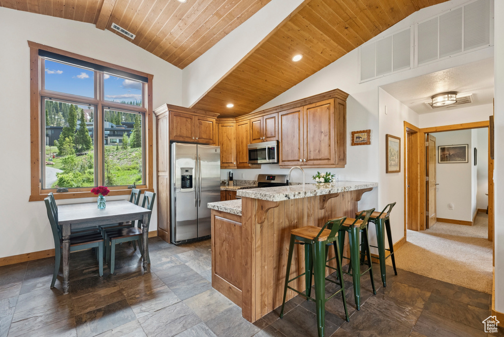 Kitchen with appliances with stainless steel finishes, vaulted ceiling, a peninsula, wood ceiling, and light stone counters