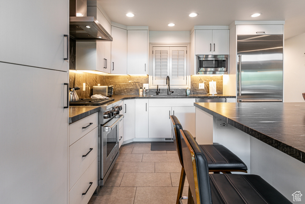 Kitchen with built in appliances, wall chimney exhaust hood, decorative backsplash, white cabinets, and recessed lighting