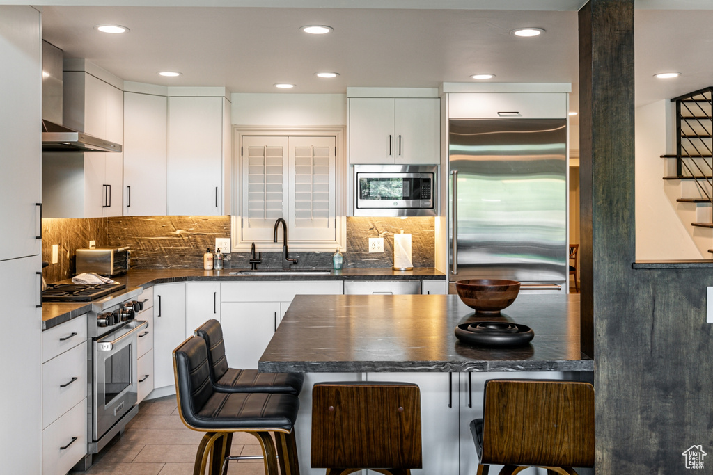 Kitchen featuring wall chimney exhaust hood, built in appliances, dark countertops, a kitchen breakfast bar, and recessed lighting
