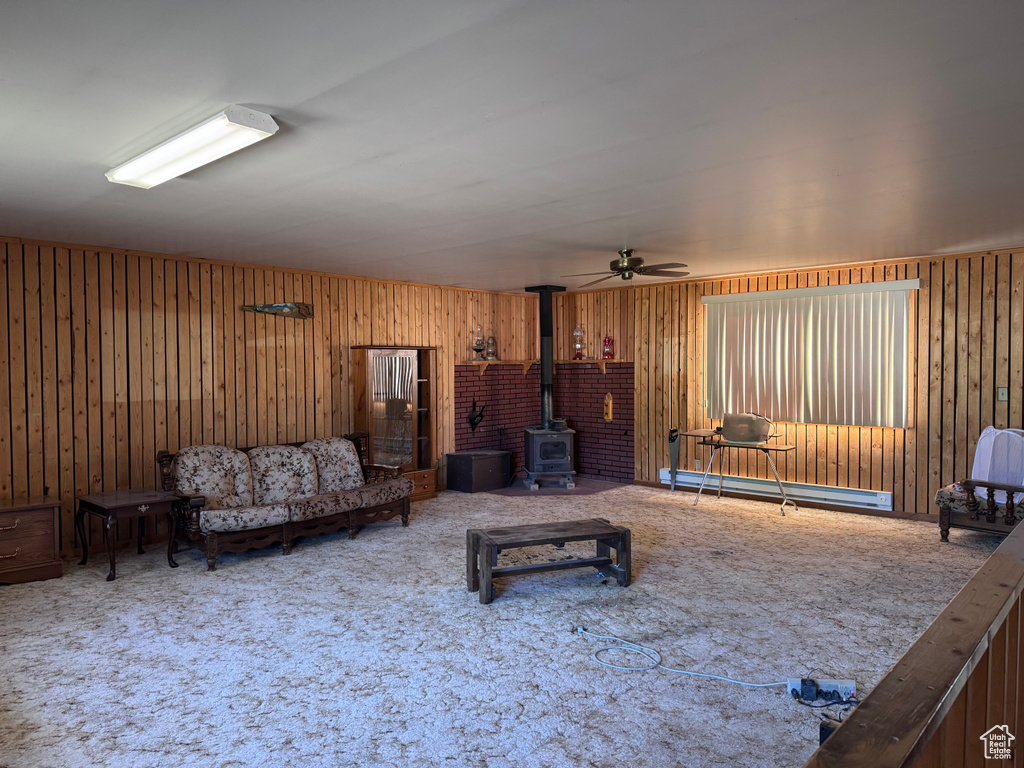 Living room featuring a baseboard radiator, a wood stove, carpet flooring, ceiling fan, and wooden walls