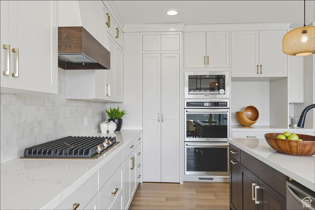 Kitchen with appliances with stainless steel finishes, wall chimney range hood, white cabinets, light wood finished floors, and decorative light fixtures
