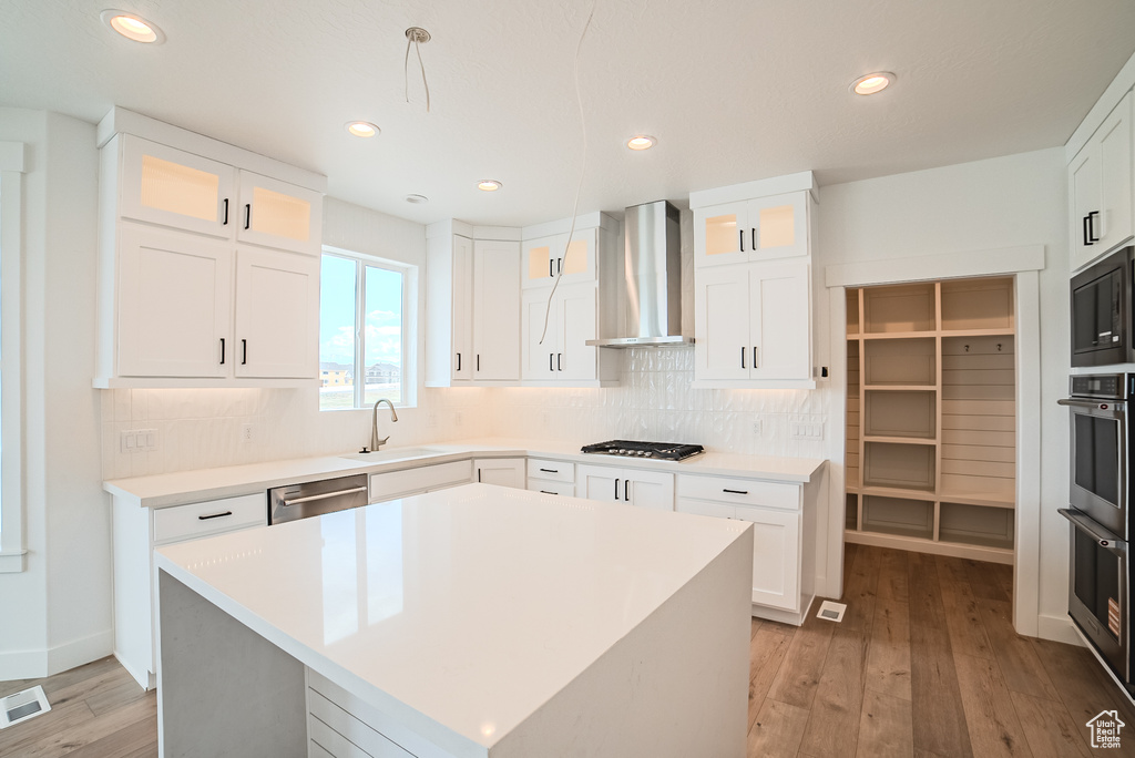 Kitchen with wall chimney exhaust hood, stainless steel appliances, backsplash, light wood-style flooring, and glass insert cabinets