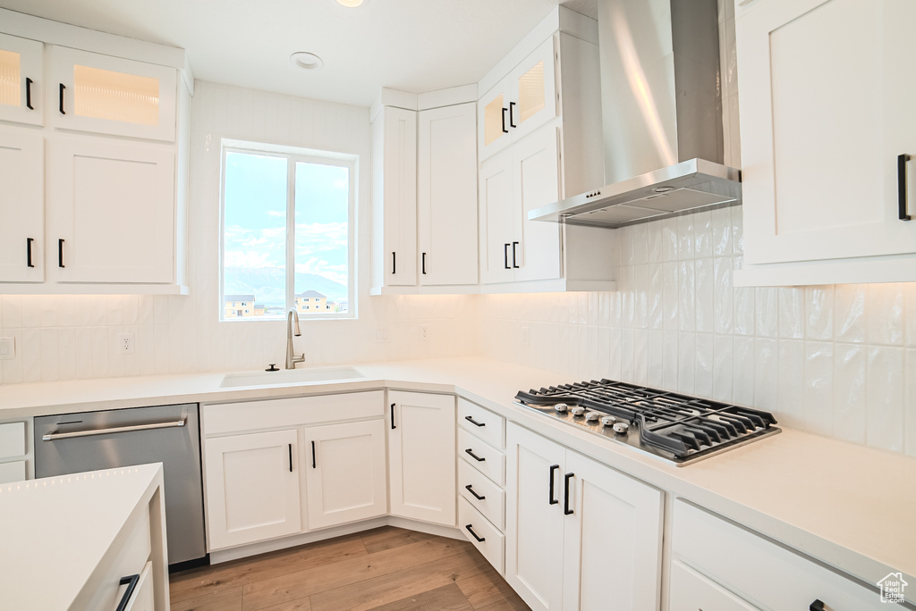 Kitchen featuring wall chimney exhaust hood, appliances with stainless steel finishes, white cabinetry, light wood finished floors, and glass insert cabinets