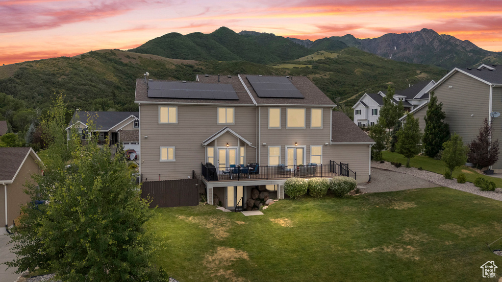 Back of property at dusk featuring solar panels, a lawn, a deck with mountain view, a chimney, and roof with shingles