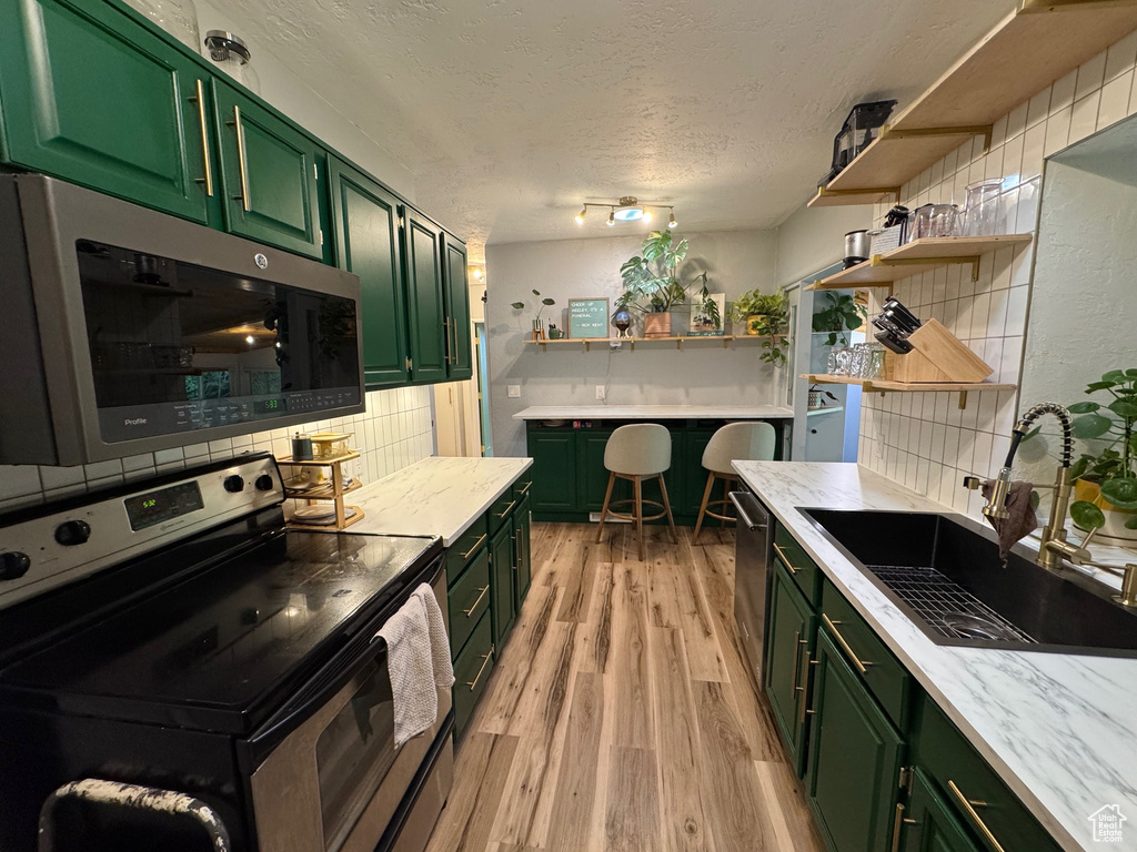 Kitchen with stainless steel appliances, green cabinets, open shelves, tasteful backsplash, and light wood finished floors