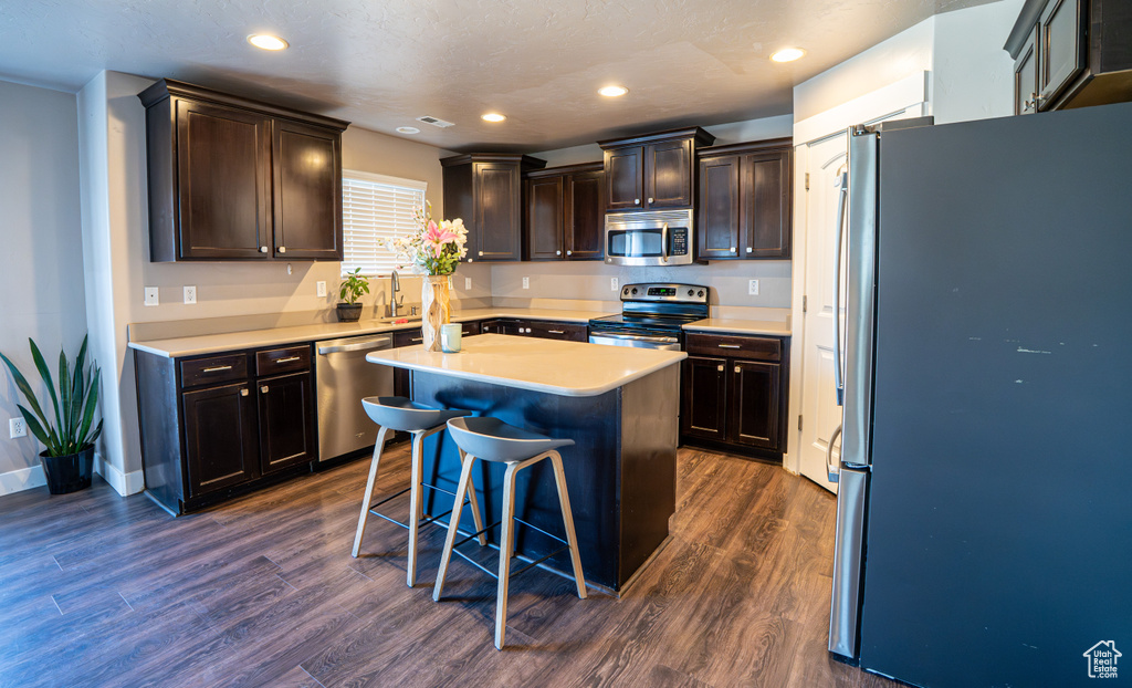 Kitchen featuring stainless steel appliances, dark wood-type flooring, light countertops, a kitchen island, and a kitchen bar