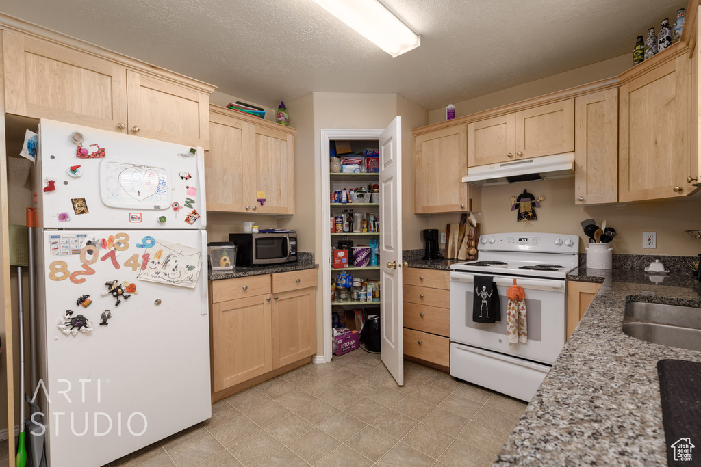 Kitchen with white appliances, light brown cabinets, under cabinet range hood, and dark stone counters