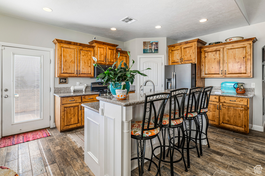 Kitchen with appliances with stainless steel finishes, brown cabinets, an island with sink, dark wood-type flooring, and recessed lighting