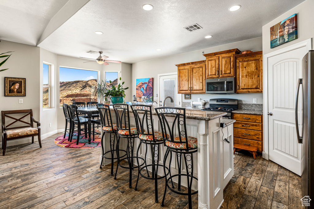 Kitchen featuring stainless steel appliances, brown cabinetry, dark wood-style floors, an island with sink, and a breakfast bar