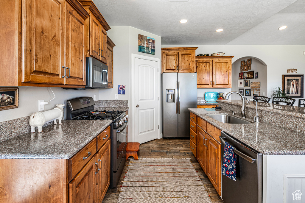 Kitchen with stainless steel appliances, brown cabinets, a center island with sink, dark stone counters, and recessed lighting