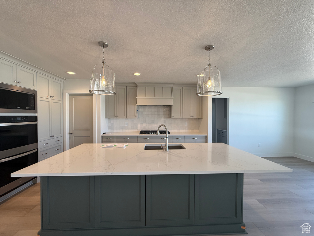 Kitchen featuring stainless steel appliances, custom exhaust hood, gray cabinets, backsplash, and a kitchen island with sink