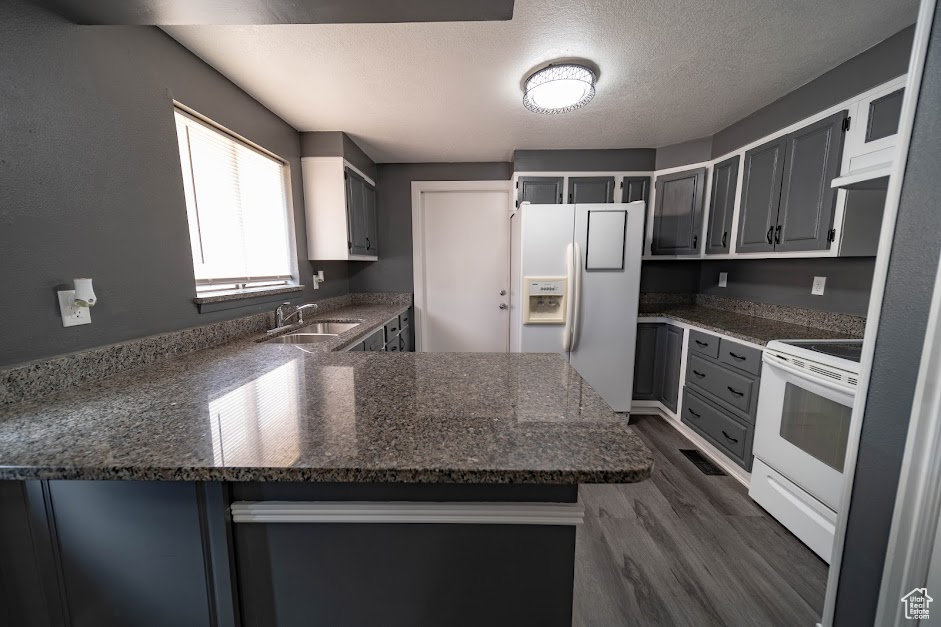Kitchen featuring white appliances, a peninsula, dark wood-type flooring, a textured ceiling, and dark stone countertops