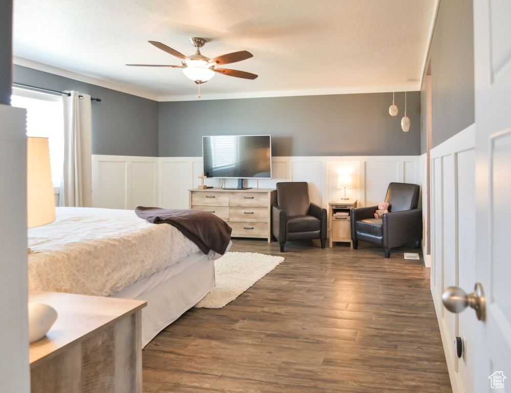 Bedroom featuring a decorative wall, a wainscoted wall, dark wood-type flooring, a ceiling fan, and ornamental molding
