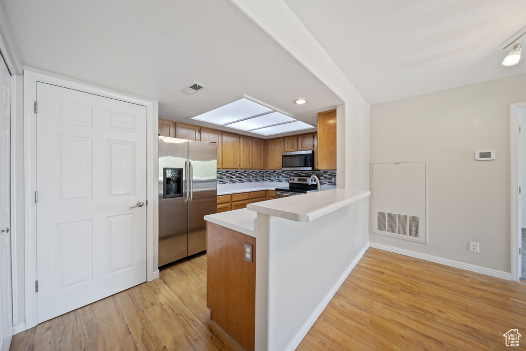 Kitchen with appliances with stainless steel finishes, light wood-type flooring, brown cabinetry, a peninsula, and recessed lighting