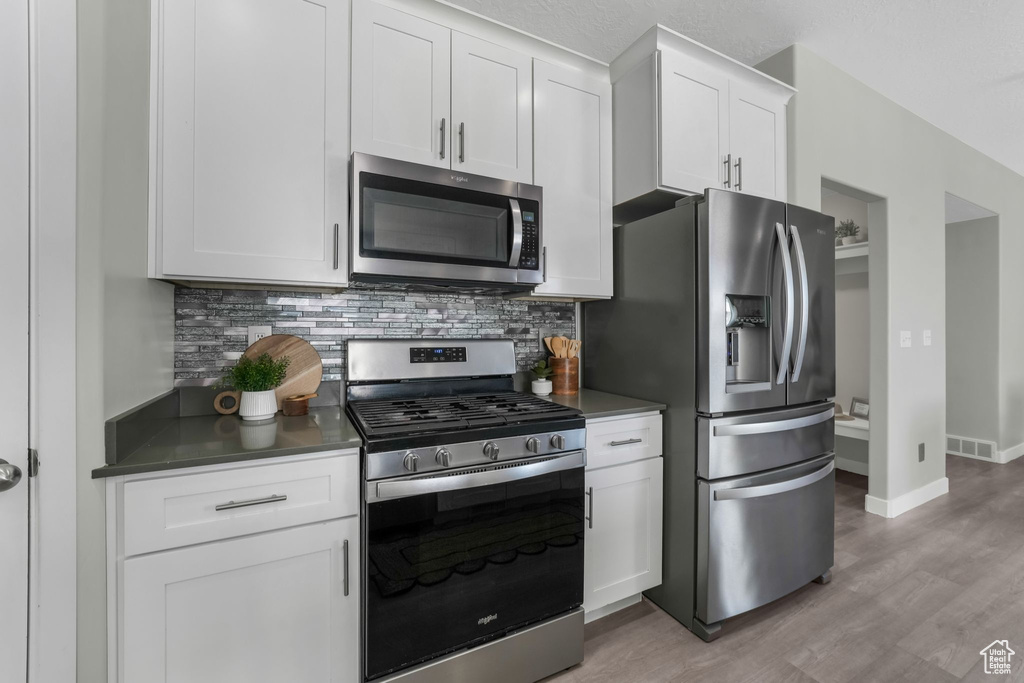 Kitchen with stainless steel appliances, decorative backsplash, light wood-style flooring, white cabinets, and dark countertops