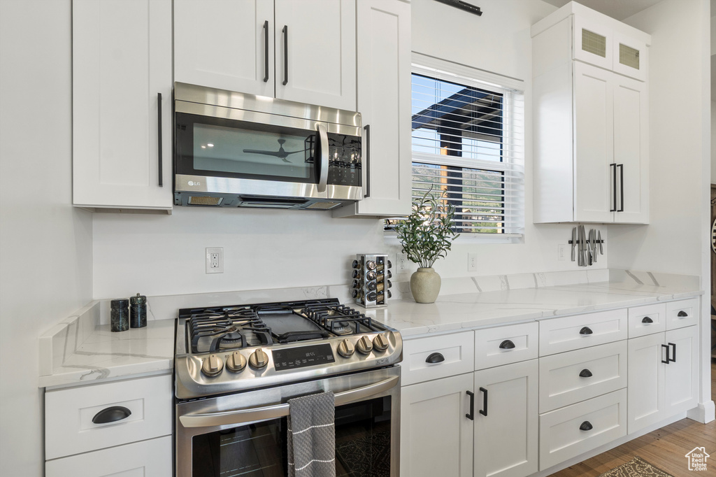 Kitchen with appliances with stainless steel finishes, white cabinets, light stone counters, wood finished floors, and glass insert cabinets