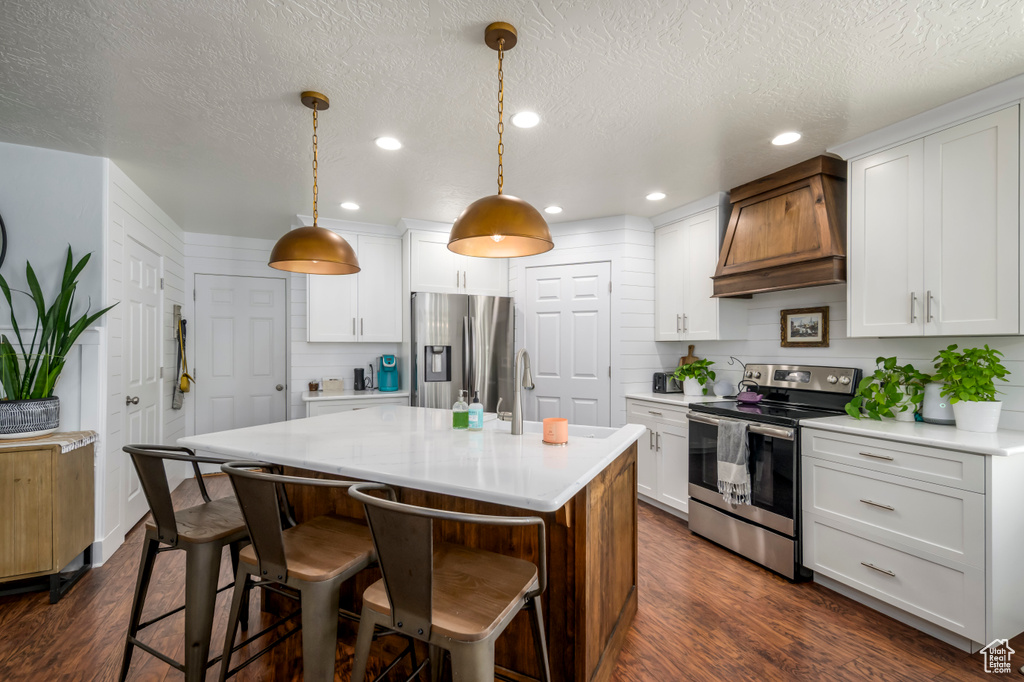 Kitchen with appliances with stainless steel finishes, premium range hood, light countertops, dark wood-style floors, and a breakfast bar
