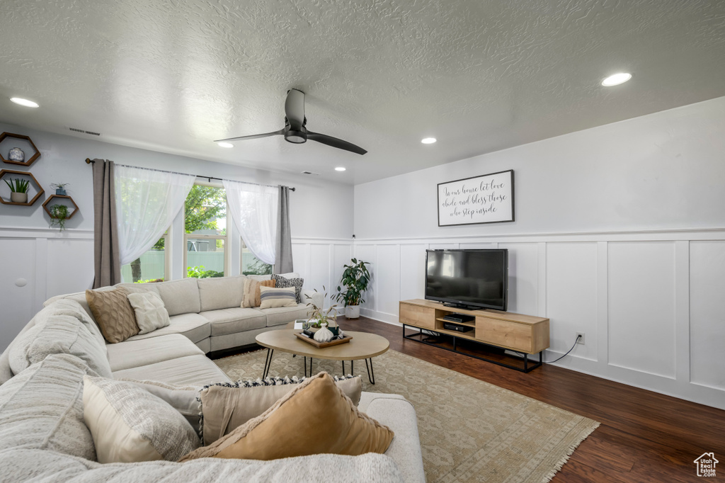 Living area featuring a decorative wall, ceiling fan, wainscoting, a textured ceiling, and dark wood-type flooring