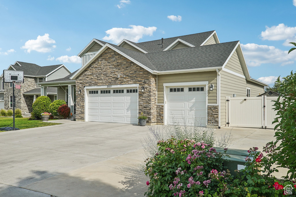 View of front of property featuring stone siding, an attached garage, concrete driveway, a gate, and roof with shingles