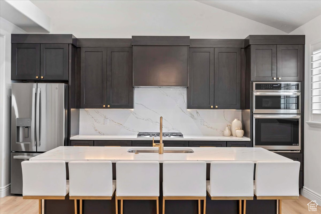 Kitchen featuring appliances with stainless steel finishes, backsplash, lofted ceiling, light wood-style floors, and a kitchen bar