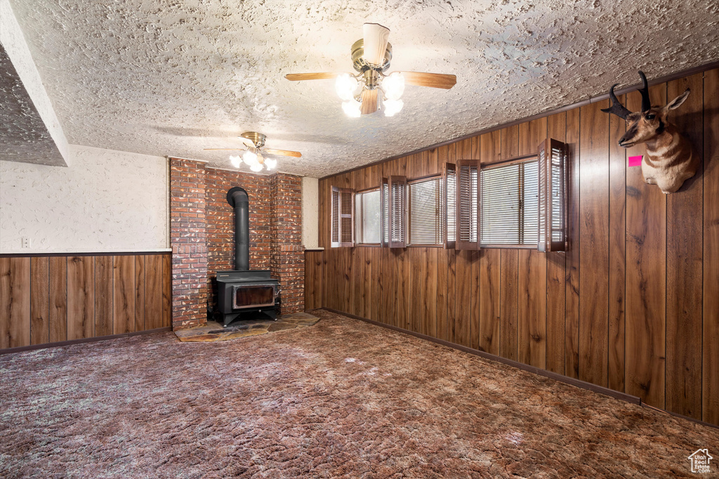 Unfurnished living room with wooden walls, a wood stove, a ceiling fan, a textured ceiling, and carpet floors