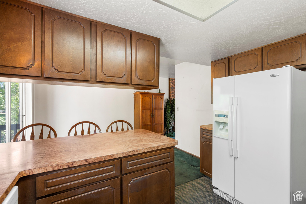 Kitchen with white fridge with ice dispenser, dark carpet, a textured ceiling, light countertops, and brown cabinets