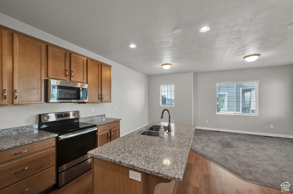 Kitchen with stainless steel appliances, brown cabinetry, an island with sink, light stone counters, and open floor plan