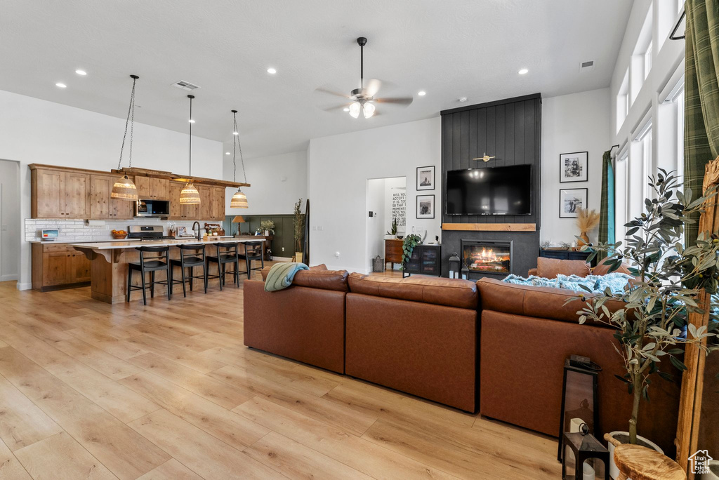 Living room with ceiling fan, recessed lighting, a large fireplace, light wood-style flooring, and a towering ceiling