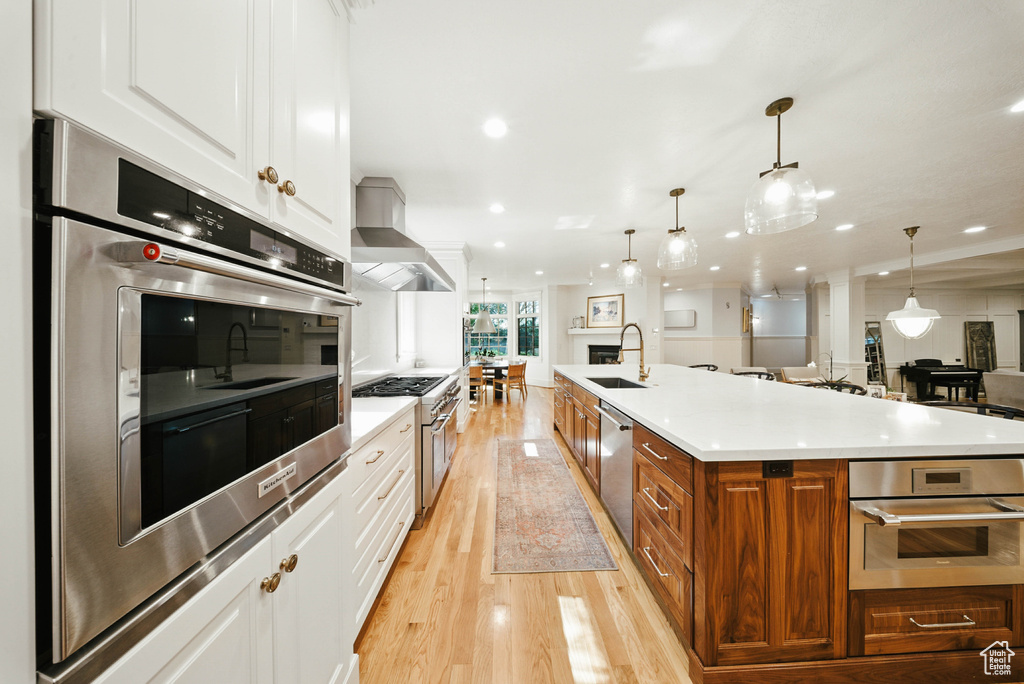Kitchen featuring wall chimney exhaust hood, stainless steel appliances, light wood finished floors, brown cabinets, and white cabinetry