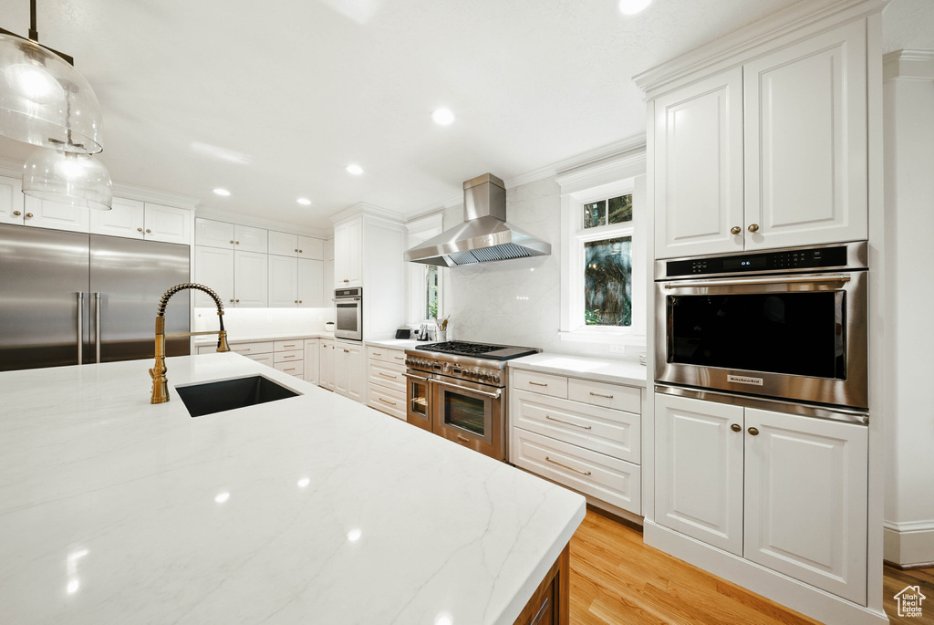 Kitchen with premium appliances, wall chimney exhaust hood, light wood finished floors, white cabinets, and recessed lighting