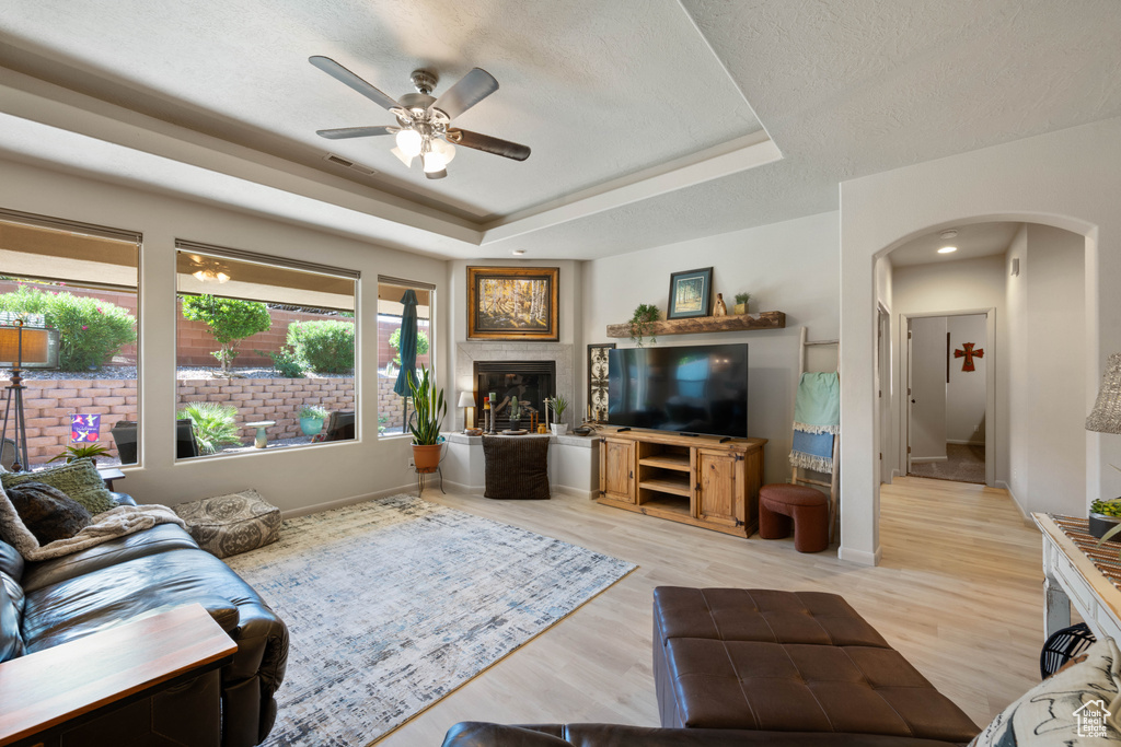 Living room with arched walkways, a raised ceiling, a ceiling fan, light wood finished floors, and a fireplace