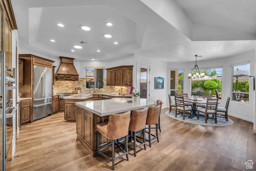 Kitchen with stainless steel appliances, premium range hood, a center island with sink, light wood-style flooring, and recessed lighting