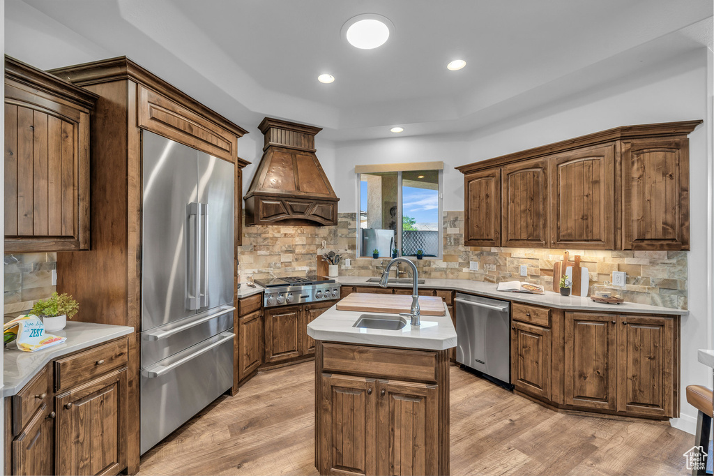 Kitchen with stainless steel appliances, custom range hood, a center island with sink, light wood-type flooring, and decorative backsplash