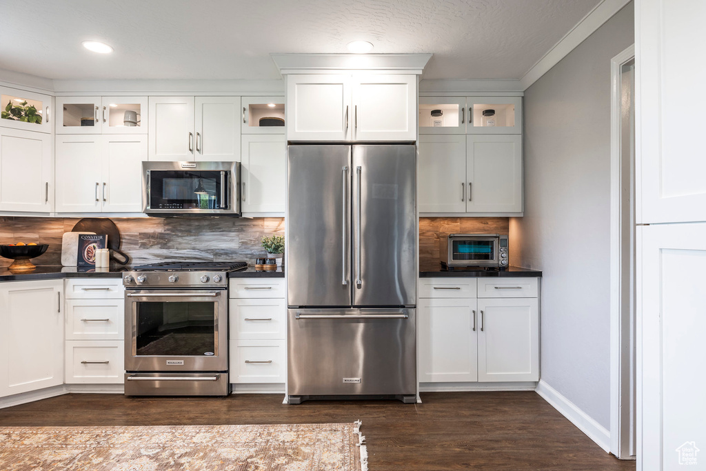 Kitchen with appliances with stainless steel finishes, dark countertops, dark wood-type flooring, tasteful backsplash, and glass insert cabinets