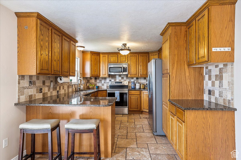 Kitchen with appliances with stainless steel finishes, a peninsula, brown cabinets, stone tile flooring, and tasteful backsplash